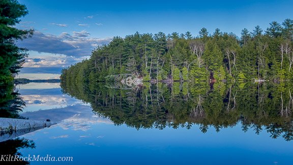 A beautiful calm, blue lake with the island and a fallen birch tree in the foreground. This landscape shot was taken as part of the filming of the 'Landscape Theatre - Lakefront' series by KiltRockMedia.com.